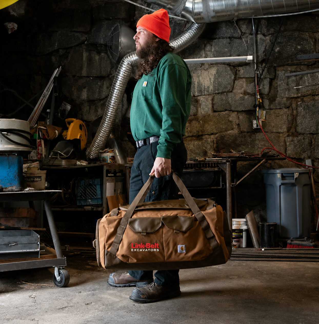 A man wearing a green sweatshirt, dark pants, and an orange beanie carries a large brown duffel bag with red and white 'LinkBait Excavators' branding in a workshop setting.